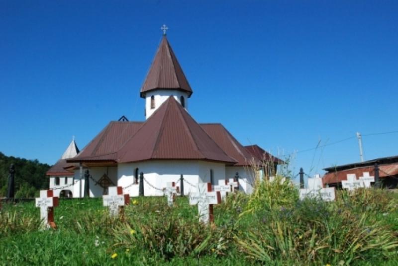 Manastirea Inaltarea Domnului Muntele Rece, o biserica alba cu acoperis rosu, inconjurata de cruci si vegetatie, situata intr-o zona rurala.