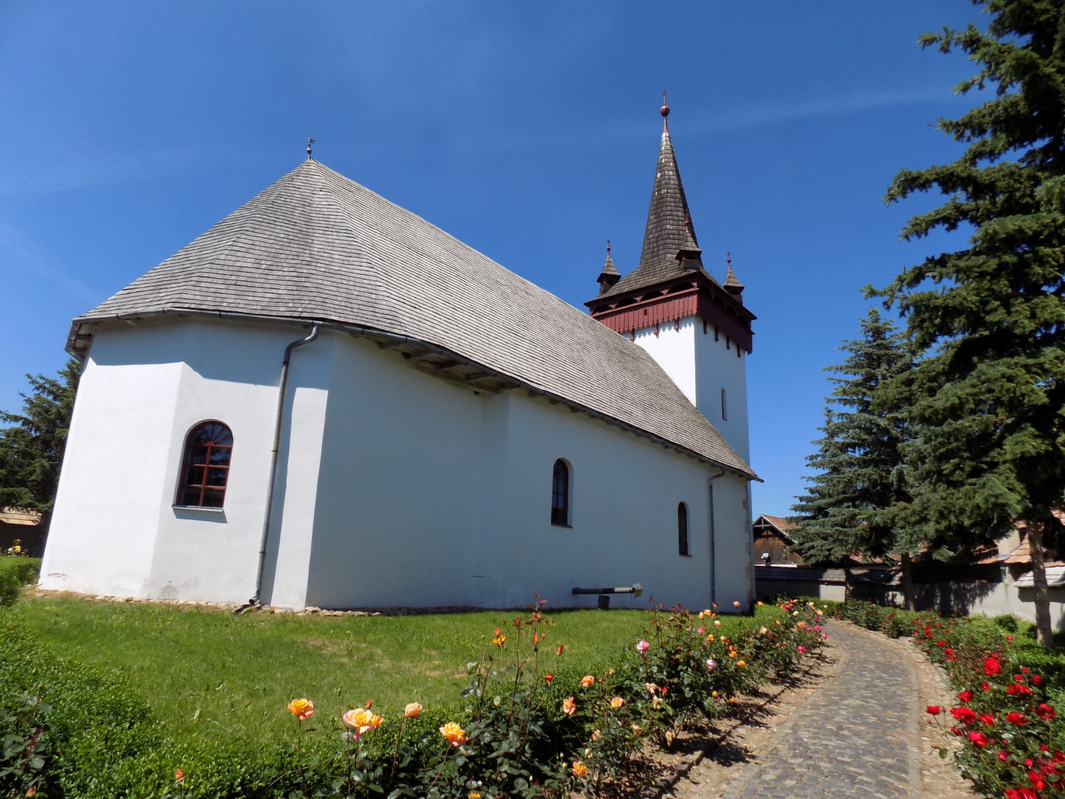 Biserica Reformat-Calvina Sancraiu, o cladire alba cu acoperis de sindrila si turn inalt, inconjurata de un drum pavat si flori colorate.